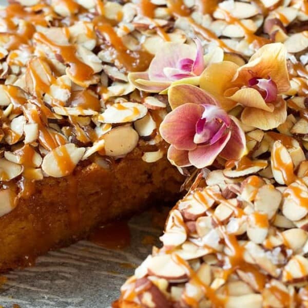 A round cake topped with sliced almonds, caramel drizzle, and two pink orchids. One slice is missing, revealing a moist, golden interior. The cake is presented on a subtle, patterned surface.