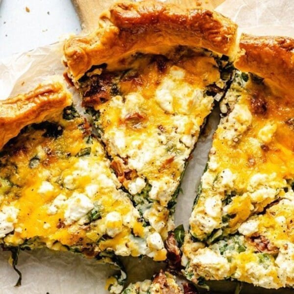 A close-up of a baked quiche with a golden crust, topped with melted cheese and herbs, sliced into wedges on a wooden surface.