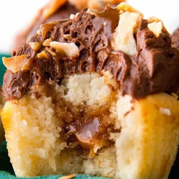 A close-up of a vanilla cupcake with chocolate frosting, chopped nuts, and a bite showing caramel filling inside.