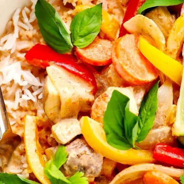 A bowl of rice topped with colorful curry containing sliced bell peppers, mushrooms, carrots, and fresh basil leaves. Lime wedges garnish the dish, and a spoon is placed on the side.