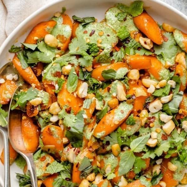A white bowl of salad with sliced carrots, fresh herbs, chopped nuts, creamy green dressing, and two serving spoons on the side.