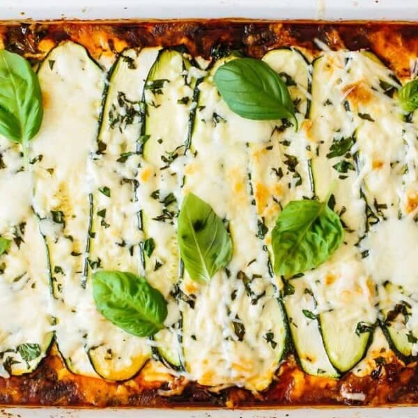 A baked casserole dish of zucchini lasagna with layers of ground beef, topped with melted cheese, chopped herbs, and basil leaves.