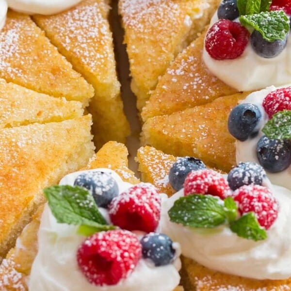 Sliced round sponge cake topped with whipped cream, blueberries, raspberries, mint leaves, and powdered sugar.