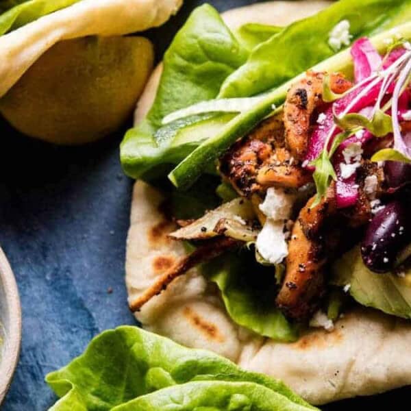 The image shows a grilled chicken wrap containing lettuce, avocado, olives, feta cheese, and pickled onions on flatbread. It is accompanied by a side of green sauce.