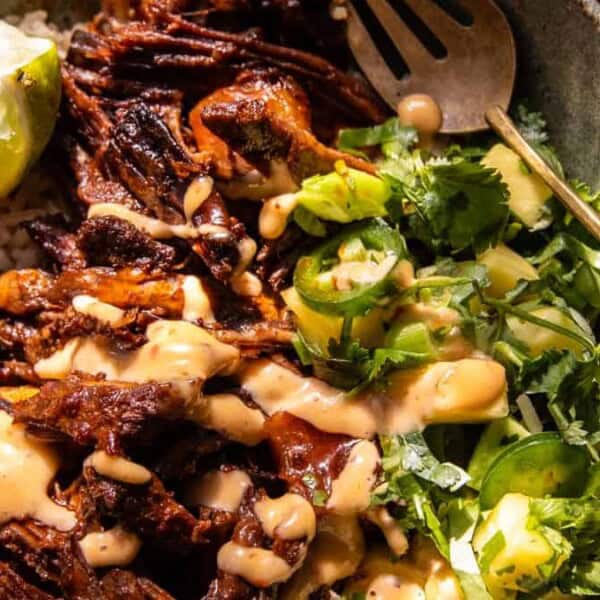 A bowl containing shredded beef, rice, creamy sauce, sliced jalapeños, cilantro, a halved lime, and a gold fork.