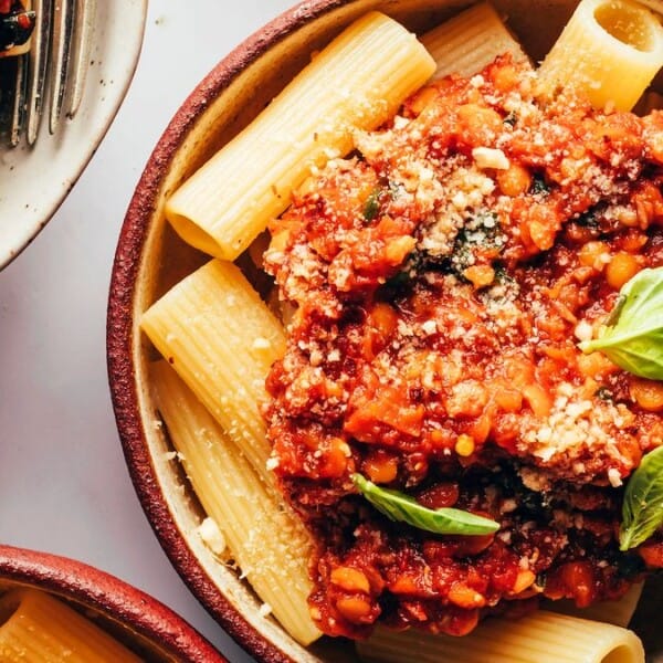 A bowl of rigatoni pasta with tomato-based lentil sauce, garnished with grated cheese and fresh basil leaves.