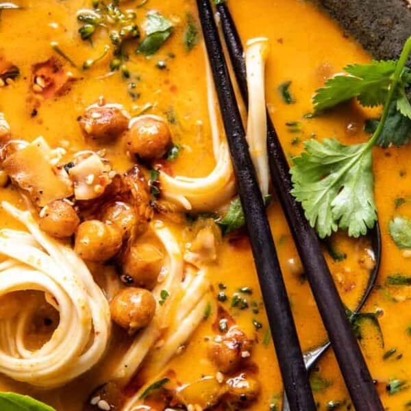 A bowl of creamy orange soup with noodles, roasted chickpeas, fresh herbs, and chopsticks resting on the edge.