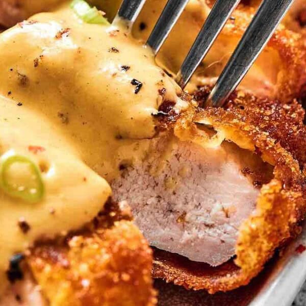 Close-up of breaded chicken breast topped with creamy sauce and sliced green onions, with a fork cutting into the meat.