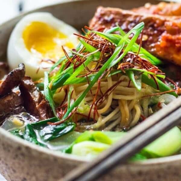 A bowl of ramen with noodles, sliced green onions, half a boiled egg, leafy greens, mushrooms, meat pieces, and chopsticks.