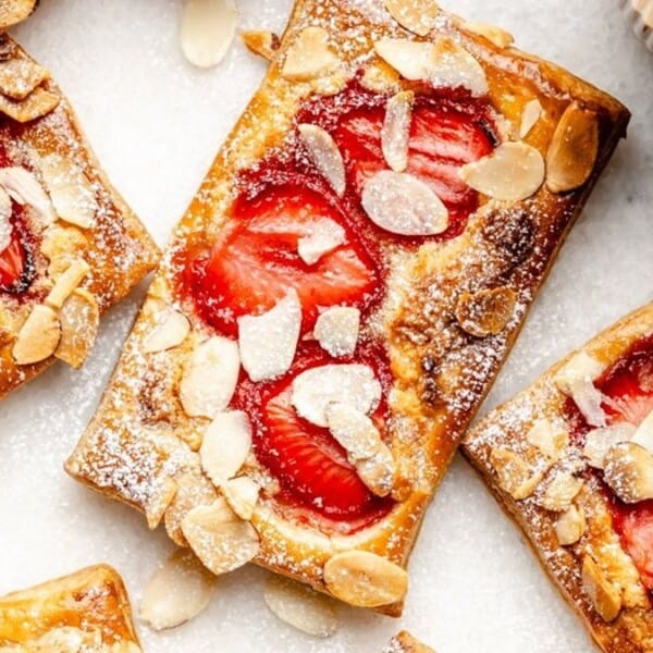 Rectangular pastries topped with sliced strawberries, almond flakes, and powdered sugar are arranged on a white surface. A cup and a whole strawberry are also partially visible in the corner.
