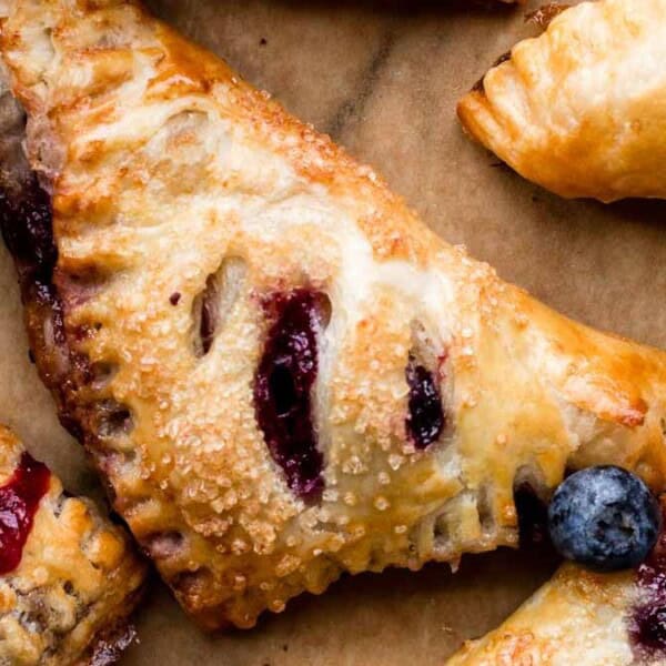 Golden-brown puff pastry turnovers with visible berry filling, sprinkled with sugar, are arranged on parchment paper beside fresh strawberries and blueberries.