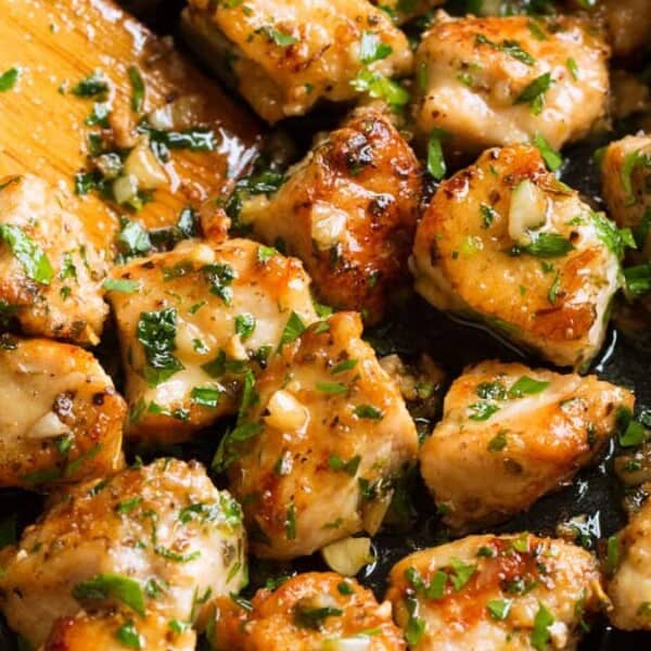 The image shows cooked chicken pieces with herbs and garlic in a pan, accompanied by a wooden spatula.