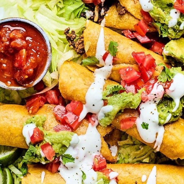 A plate of crispy taquitos topped with guacamole, diced tomatoes, and a drizzle of white sauce, served on a bed of shredded lettuce with lime wedges and a side of salsa.