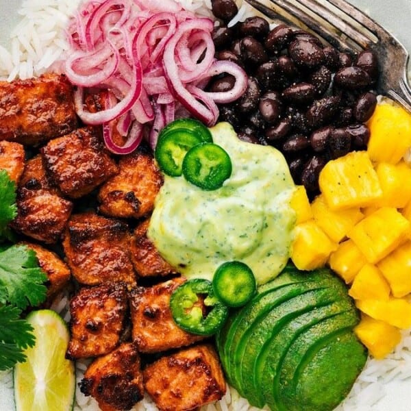 The image features a bowl containing rice, grilled meat, sliced avocado, diced pineapple, black beans, pickled onions, sliced jalapeños, cilantro, a lime wedge, and a creamy sauce. A fork is positioned beside the bowl.
