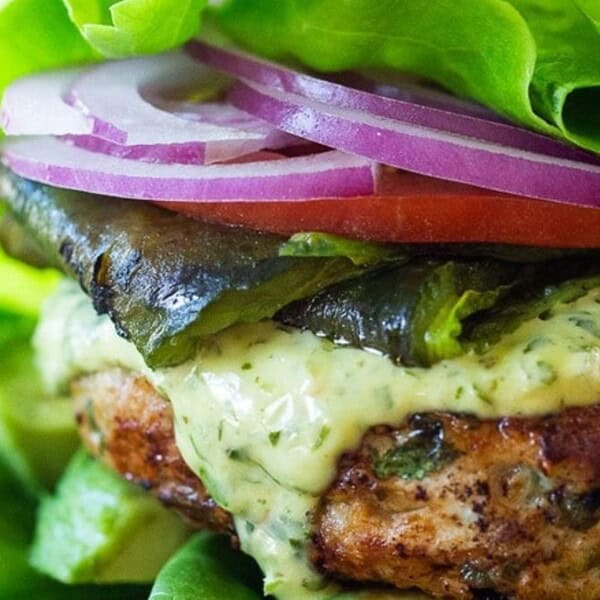 A close-up of a lettuce-wrapped burger topped with creamy sauce, roasted green pepper, sliced tomato, and red onion rings. The burger patty is grilled and surrounded by fresh green lettuce leaves.