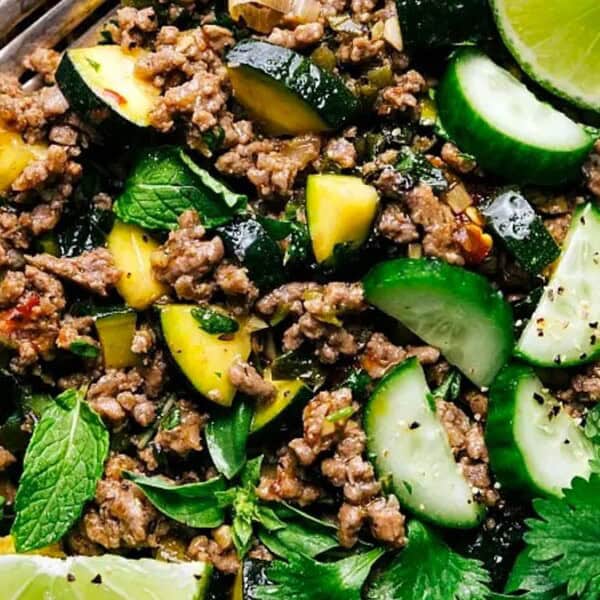 A plate of ground beef stir-fry with chopped zucchini, fresh cucumber slices, lime wedges, mint, cilantro, and white rice, served with a fork.
