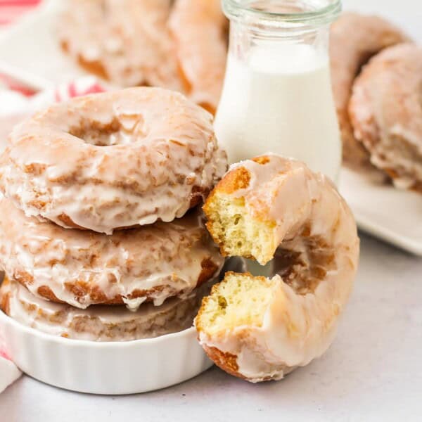A stack of glazed donuts sits in a white dish next to a glass bottle of milk. One donut has a bite taken out of it. More donuts are on a plate in the background, near a red and white striped towel.