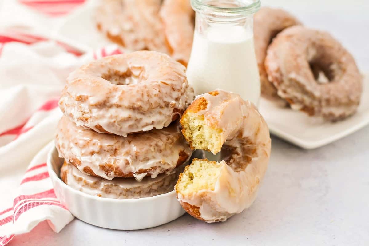 A stack of glazed donuts sits in a white dish next to a glass bottle of milk. One donut has a bite taken out of it. More donuts are on a plate in the background, near a red and white striped towel.