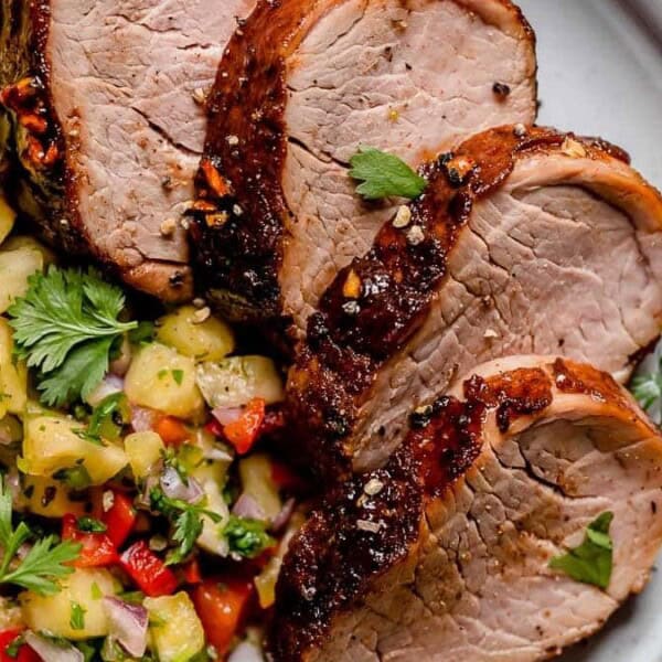 Sliced pork tenderloin with pineapple salsa featuring chopped red onion, red bell pepper, and cilantro on a white plate.