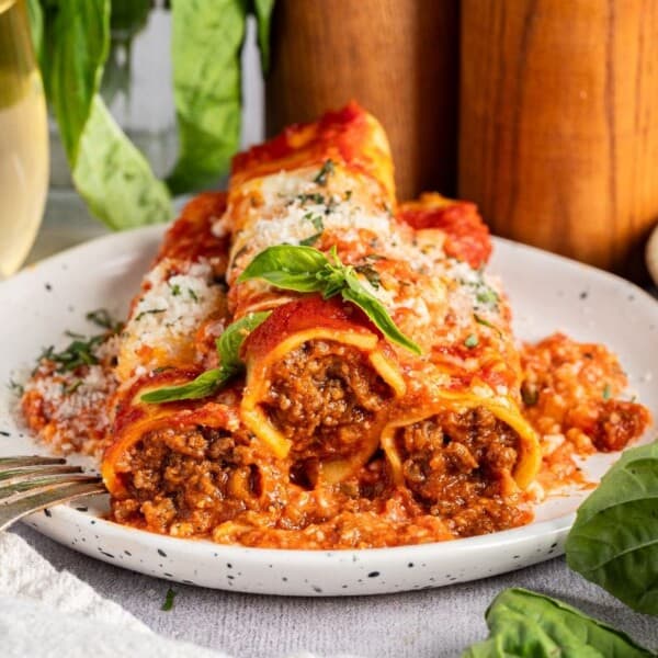 A plate of cannelloni pasta stuffed with ground meat, topped with tomato sauce, grated cheese, and fresh basil, sits beside a fork. Green herbs and a glass of white wine are in the background.