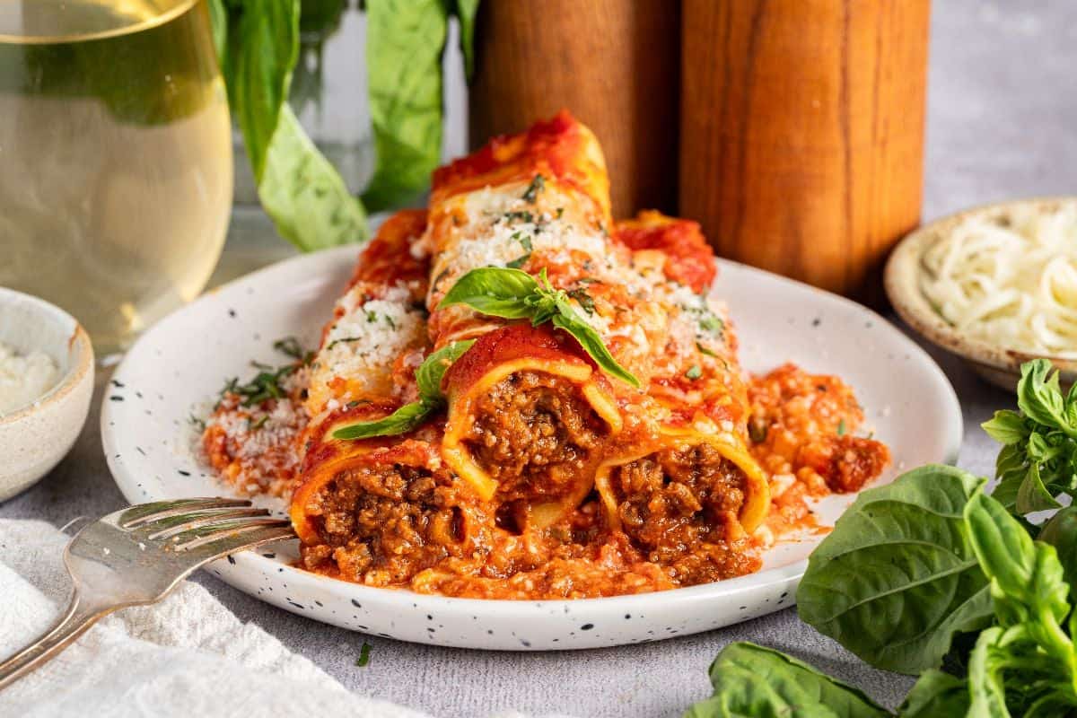 A plate of cannelloni pasta stuffed with ground meat, topped with tomato sauce, grated cheese, and fresh basil, sits beside a fork. Green herbs and a glass of white wine are in the background.