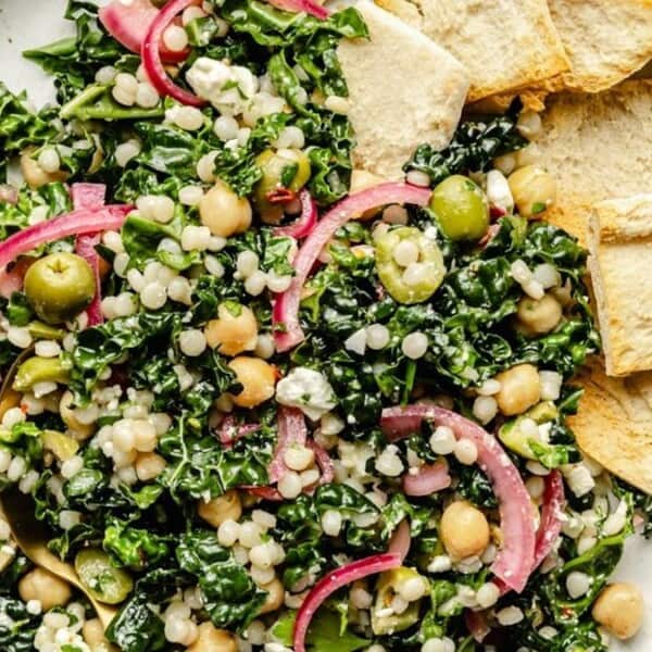 A plate with kale salad containing pearl couscous, chickpeas, green olives, pickled red onions, and feta cheese, served with pita chips on the side.