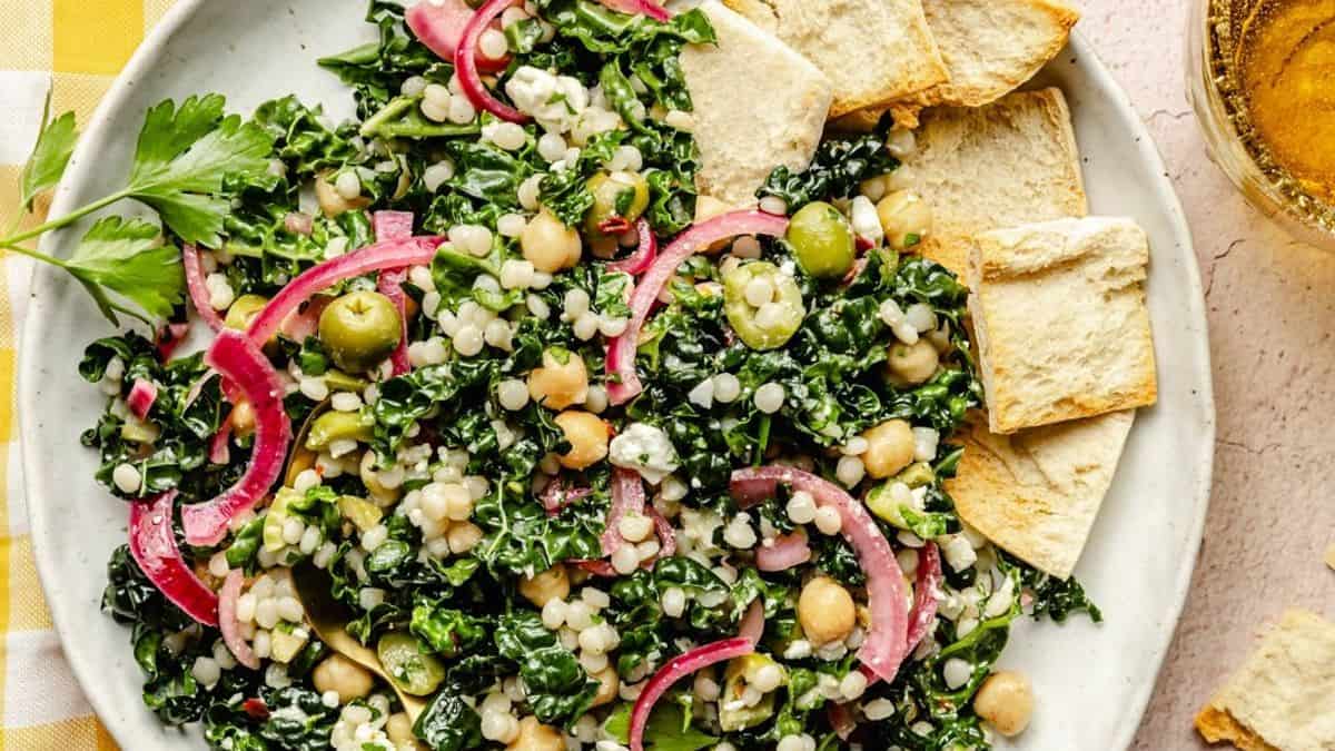 A plate with kale salad containing pearl couscous, chickpeas, green olives, pickled red onions, and feta cheese, served with pita chips on the side.