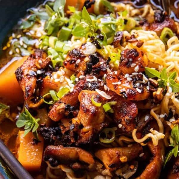 A bowl of ramen noodles topped with glazed chicken, cubed squash, green onions, sesame seeds, and fresh herbs, with a rich broth and a pair of chopsticks resting on the side.
