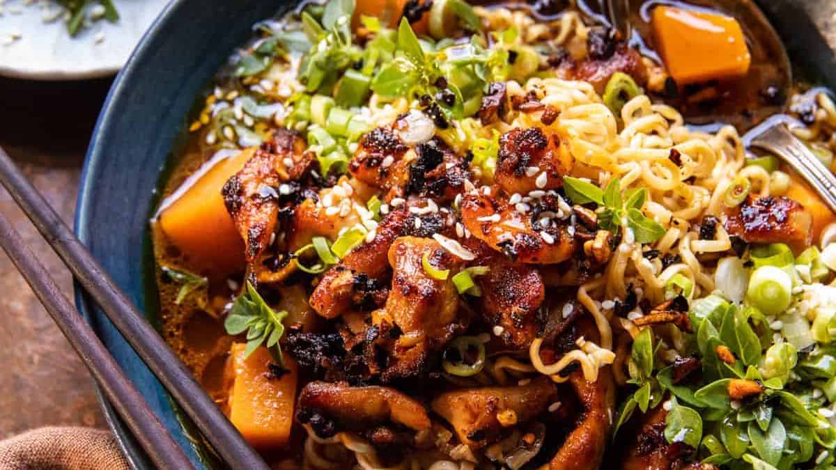 A bowl of ramen noodles topped with glazed chicken, cubed squash, green onions, sesame seeds, and fresh herbs, with a rich broth and a pair of chopsticks resting on the side.