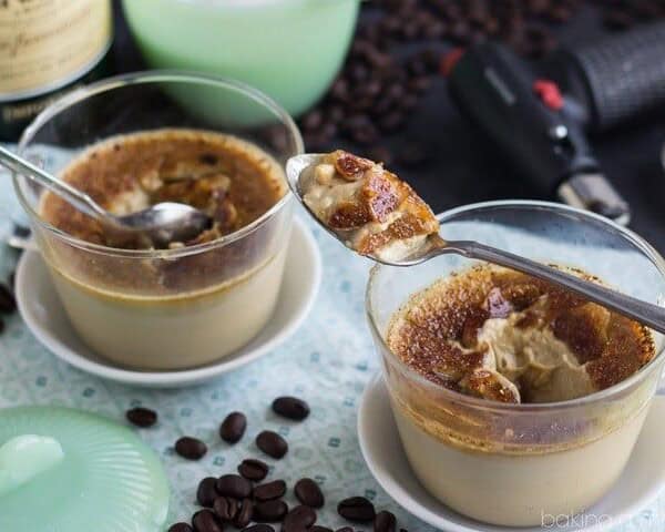 The image displays two glass cups filled with a caramel dessert that is topped with a crunchy layer. Next to the cups are spoons, and coffee beans can be seen scattered on the table along with a torch.