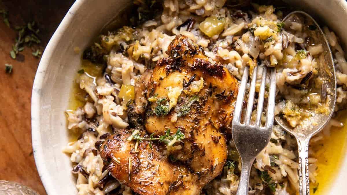 A close-up of a bowl with creamy wild rice and chicken, topped with herbs. A fork and spoon rest beside a juicy, browned chicken thigh on top of the rice. The dish has a rustic, hearty appearance.