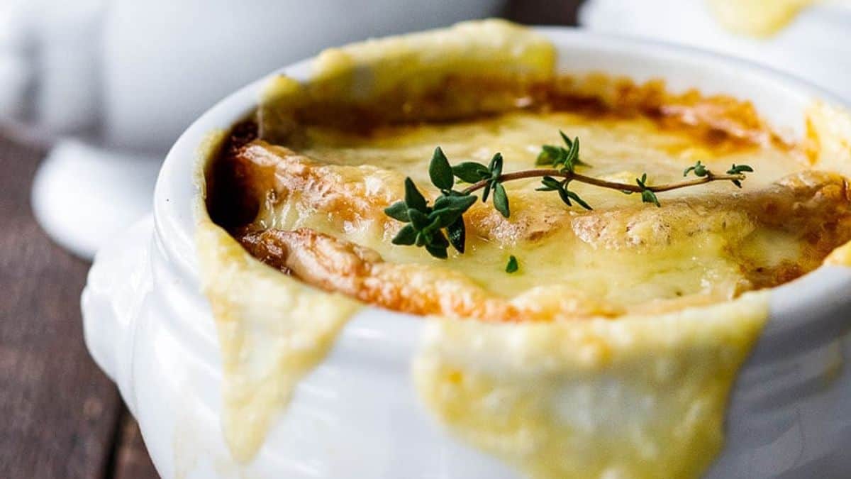 A close-up of a bowl of French onion soup topped with melted, golden-brown cheese, garnished with a sprig of fresh thyme, with cheese draping over the edge of the white bowl.