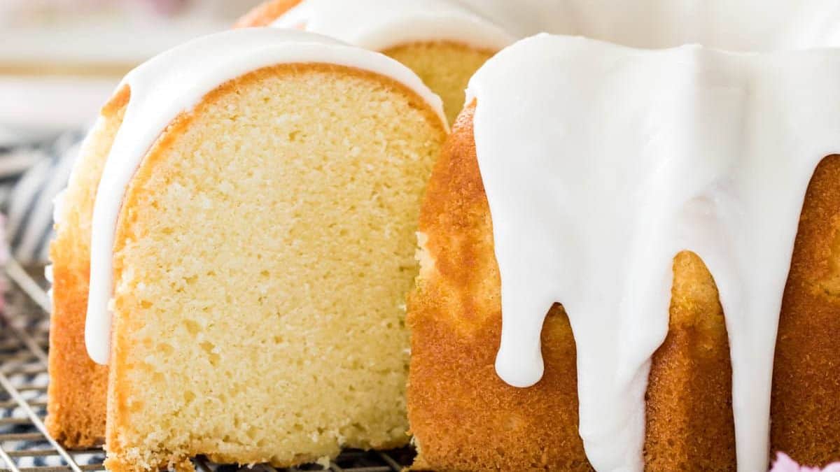 A moist bundt cake with a golden crust, topped with thick white icing that drips down the sides. A slice has been cut, revealing the soft, dense interior. The cake sits on a cooling rack.