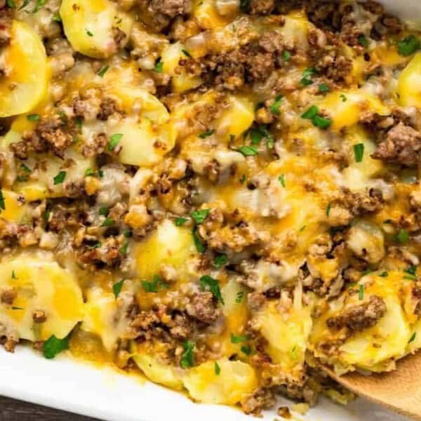 A white casserole dish with sliced potatoes, ground beef, melted cheese, and herbs. A wooden spoon scoops out a portion.