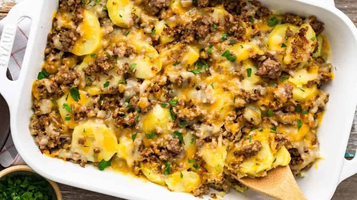 A white baking dish filled with a casserole made of sliced potatoes, ground beef, melted cheese, and topped with chopped herbs. A wooden spoon rests in the dish, ready for serving.