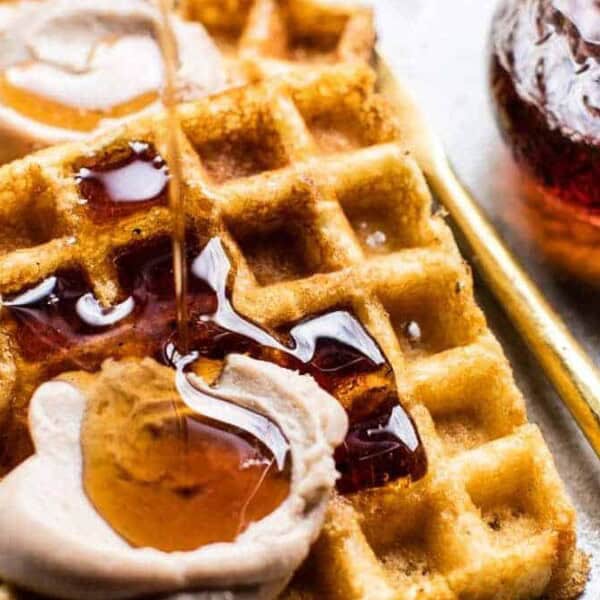 Two waffles topped with whipped cream and syrup, more syrup being poured on top. A small glass of syrup and a gold spoon beside them.