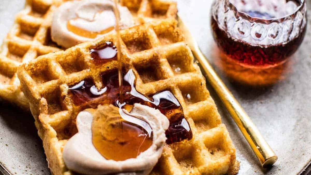 Two golden waffles topped with dollops of cream, being drizzled with syrup, next to a small glass container of syrup and a gold-colored fork on a plate.