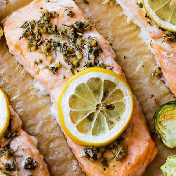 Three baked salmon fillets with lemon slices and herbs on parchment paper, served with roasted Brussels sprouts.