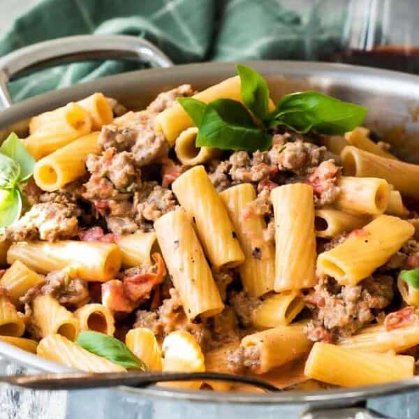 A stainless steel pan filled with rigatoni pasta, ground meat, tomatoes, and fresh basil leaves.