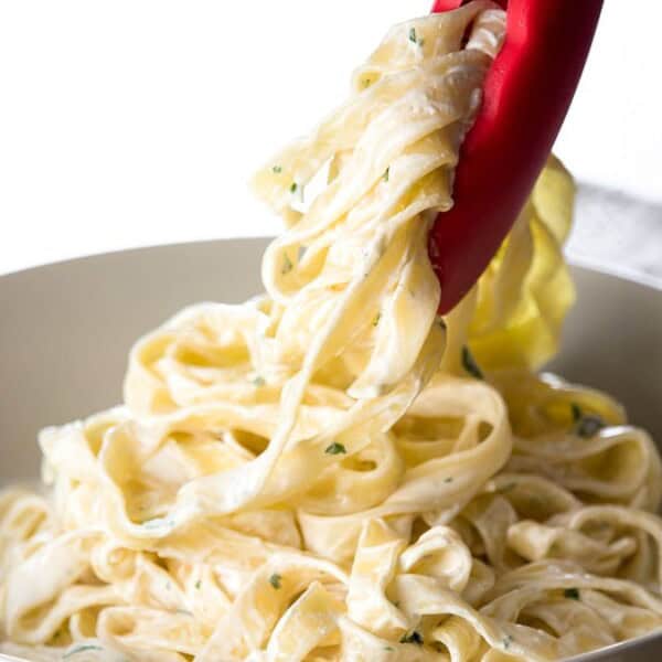 Creamy fettuccine Alfredo pasta in a white bowl, with red tongs on a light surface.