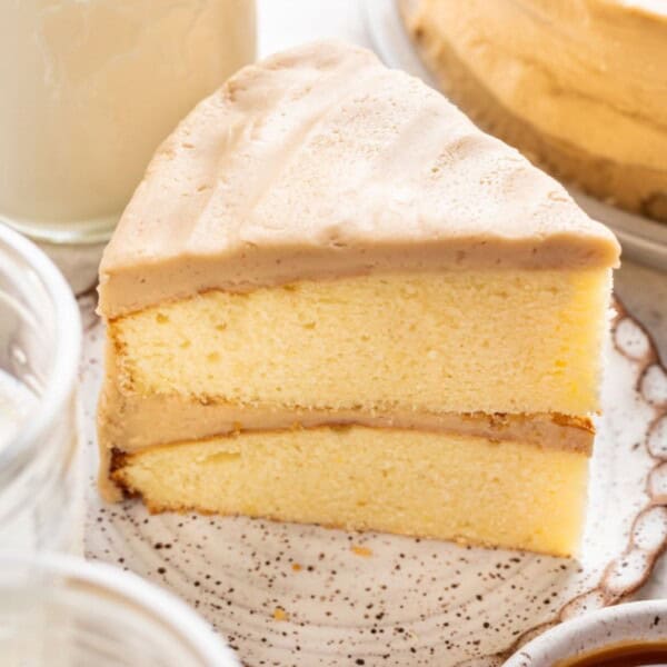 A slice of yellow layer cake with light brown frosting on a plate next to a glass of milk and several jars.