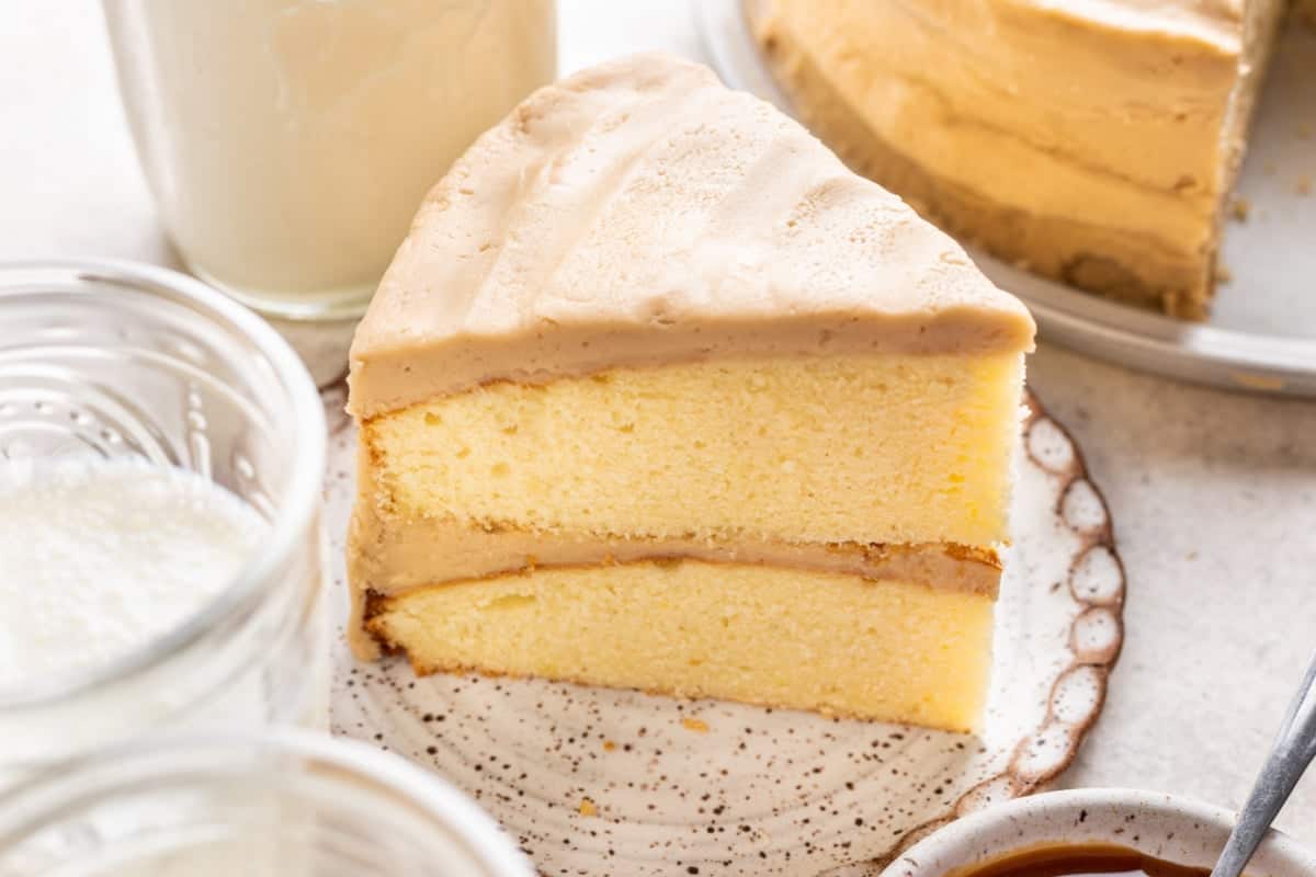 A slice of yellow layer cake with light brown frosting sits on a plate. The cake has two layers and smooth frosting on top and between the layers. Jars and a drink are visible nearby.