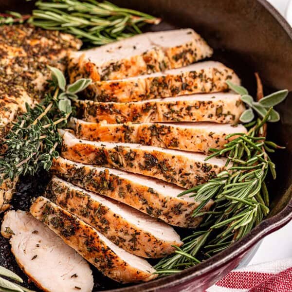 Sliced herb-seasoned turkey breast in a cast iron skillet, garnished with rosemary and thyme, with a cup of sauce on the side.