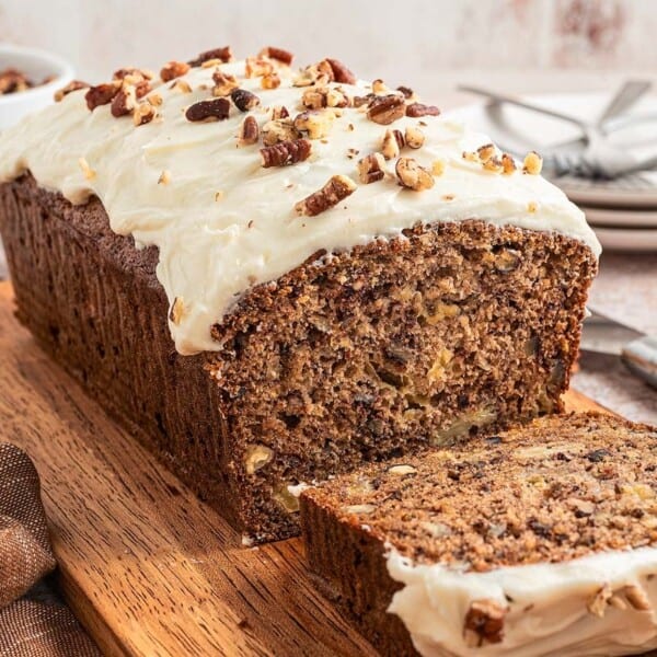 A sliced loaf of banana bread topped with creamy frosting and chopped pecans sits on a wooden board, with plates, forks, and a bowl of pecans in the background.