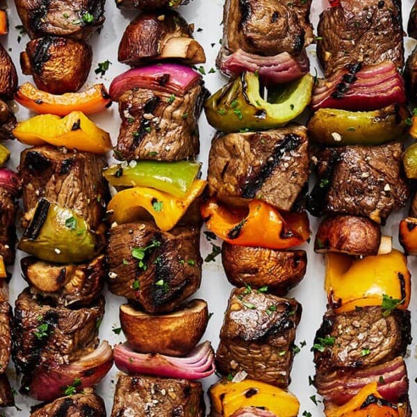 Grilled beef kebabs with mushrooms, red onions, and bell peppers in green, yellow, and orange arranged on skewers on a white tray. The kebabs are garnished with chopped parsley.