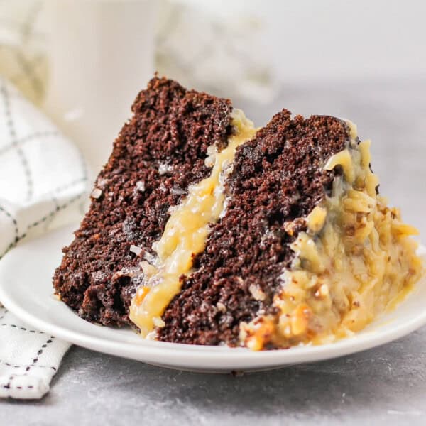 A slice of chocolate cake with coconut pecan frosting on a white plate next to a checkered cloth.