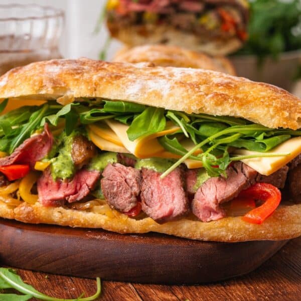 A ciabatta sandwich filled with sliced steak, melted cheese, arugula, grilled peppers, and green sauce, served on a wooden board with onion rings in the background.