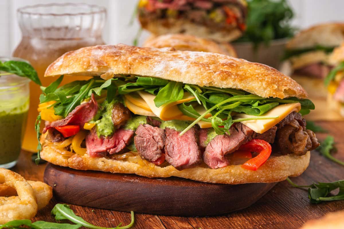 A ciabatta sandwich filled with sliced steak, melted cheese, arugula, grilled peppers, and green sauce, served on a wooden board with onion rings in the background.