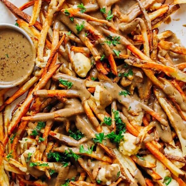 French fries topped with brown gravy, cheese curds, and chopped parsley, served on a plate with a small bowl of extra gravy on the side.