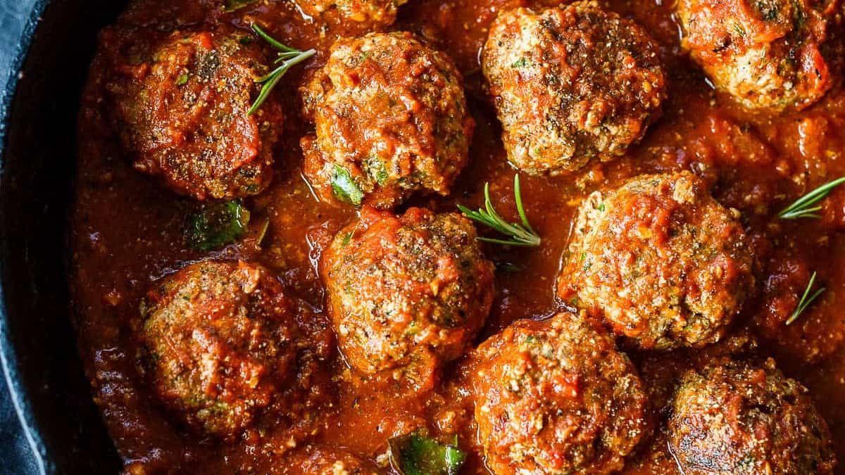 Meatballs simmering in tomato sauce, garnished with fresh rosemary sprigs.